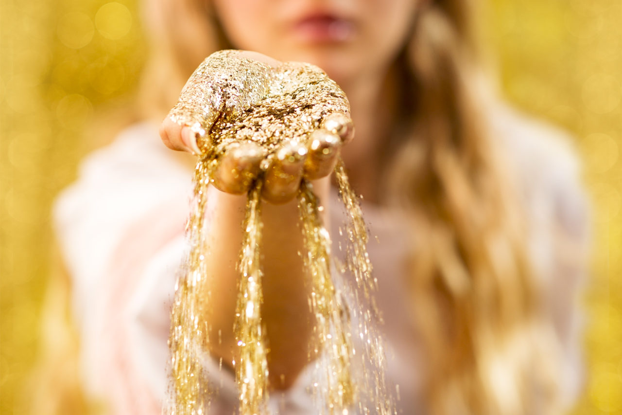 Against a very golden and sparkly backdrop, there is an outstretched hand full of glitter that spills out from between the fingers.