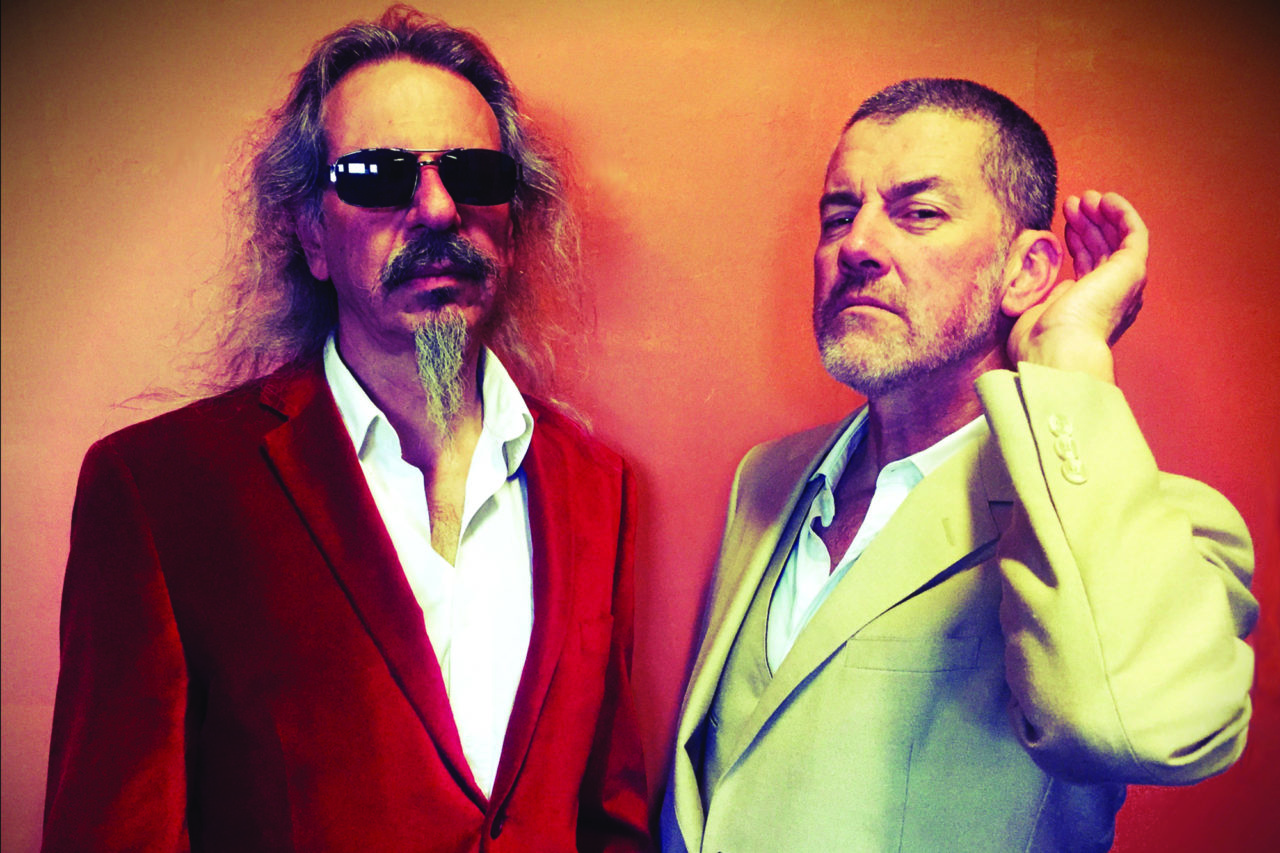 two men stand one with sunglasses on the other holding his hand to his ear