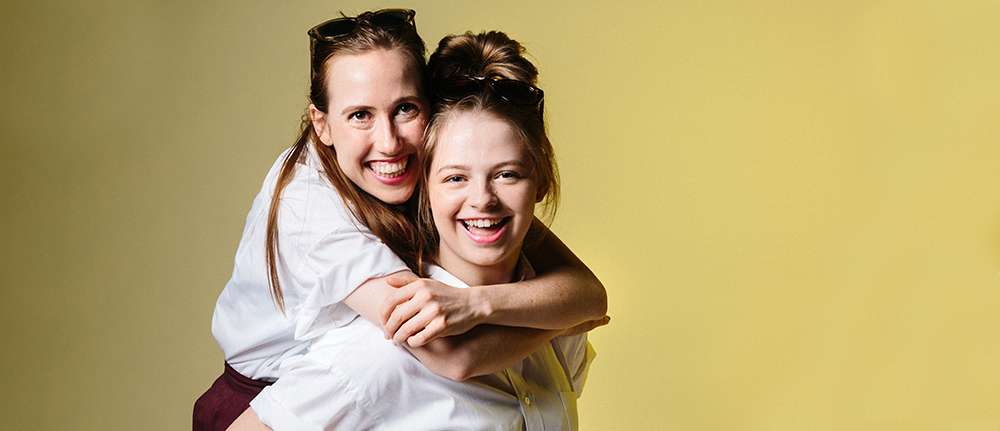 Two teenage girls in school uniform. One hugging the other from behind they both look into the camera smiling.