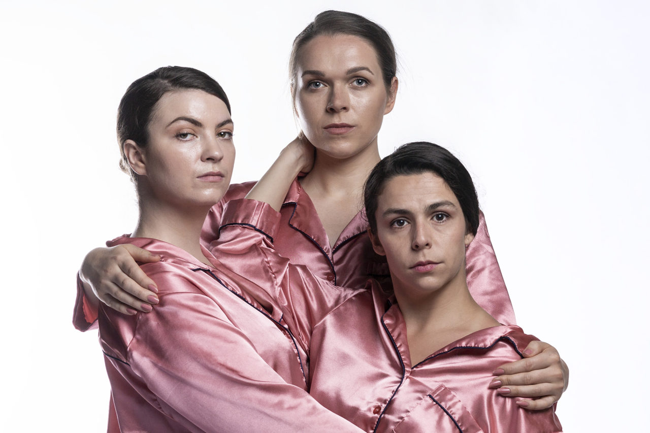 Three people in pink satin pyjamas holding each other and looking into the camera