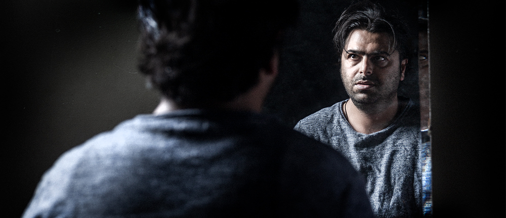 The back of a man’s head you can see his face looking back into a mirror. He has dark hair and skin and has a distressed expression on his face.