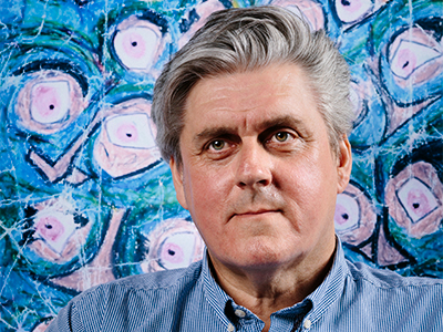 A close up of a man with grey hair smiling into the camera with a blue, pink and Green artwork background