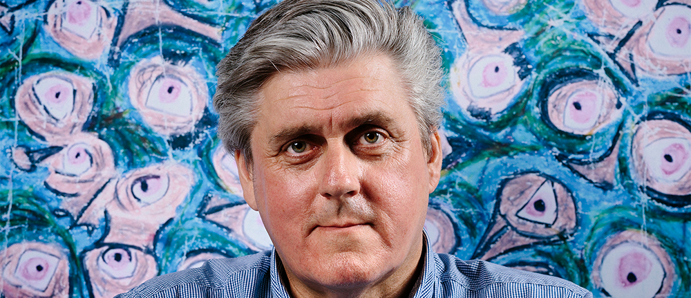 A close up of a man with grey hair smiling into the camera with a blue, pink and Green artwork background