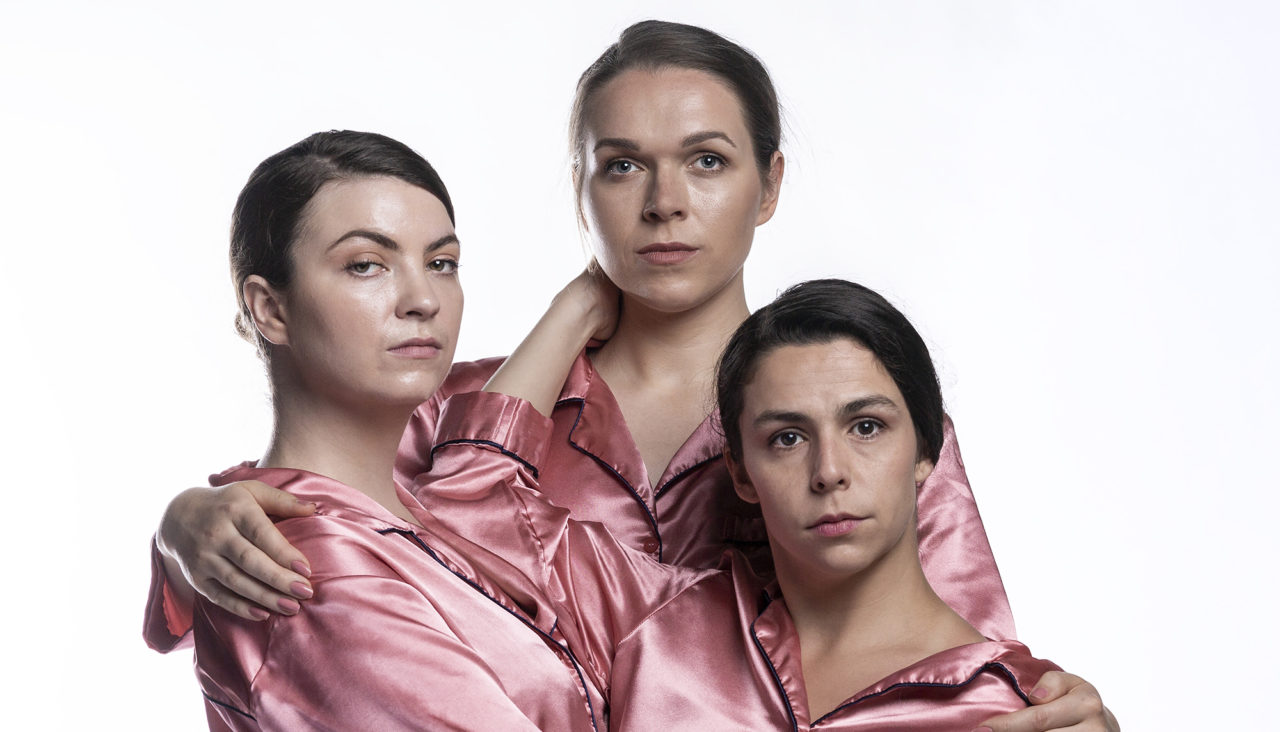 Three people in pink satin pyjamas holding each other and looking into the camera