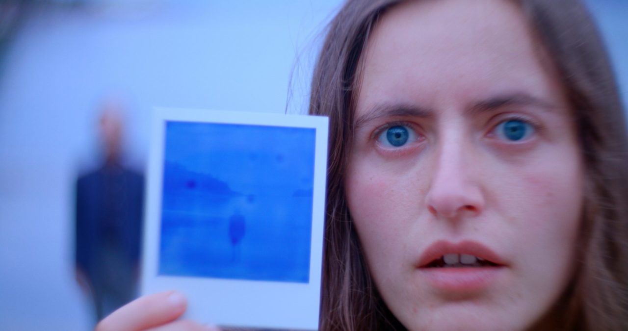 A person with fair skin and hair. They have piercing blue eyes and are holding a polaroid picture up to the camera.