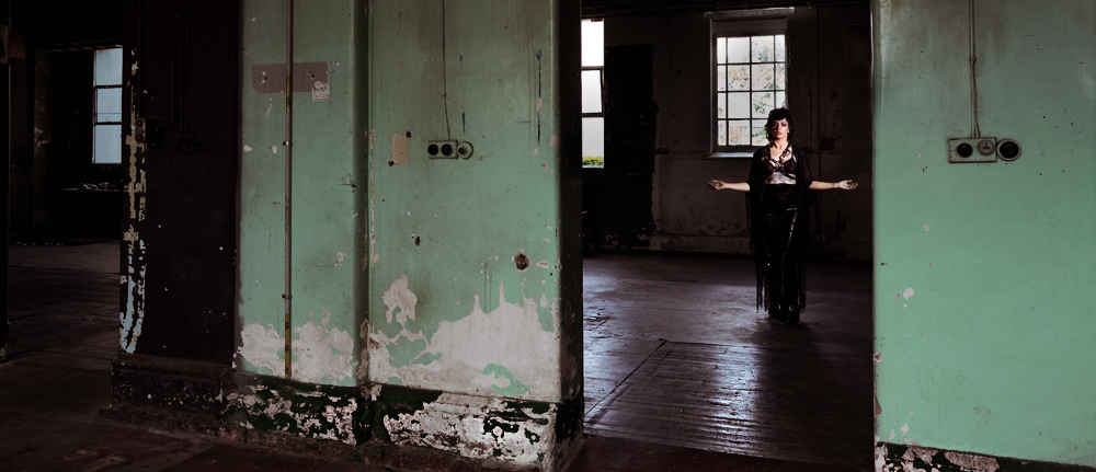 two doorways set into an old, neglected wall lead into the room of an abandoned building; in the background of one, a figure in all black can just be made out
