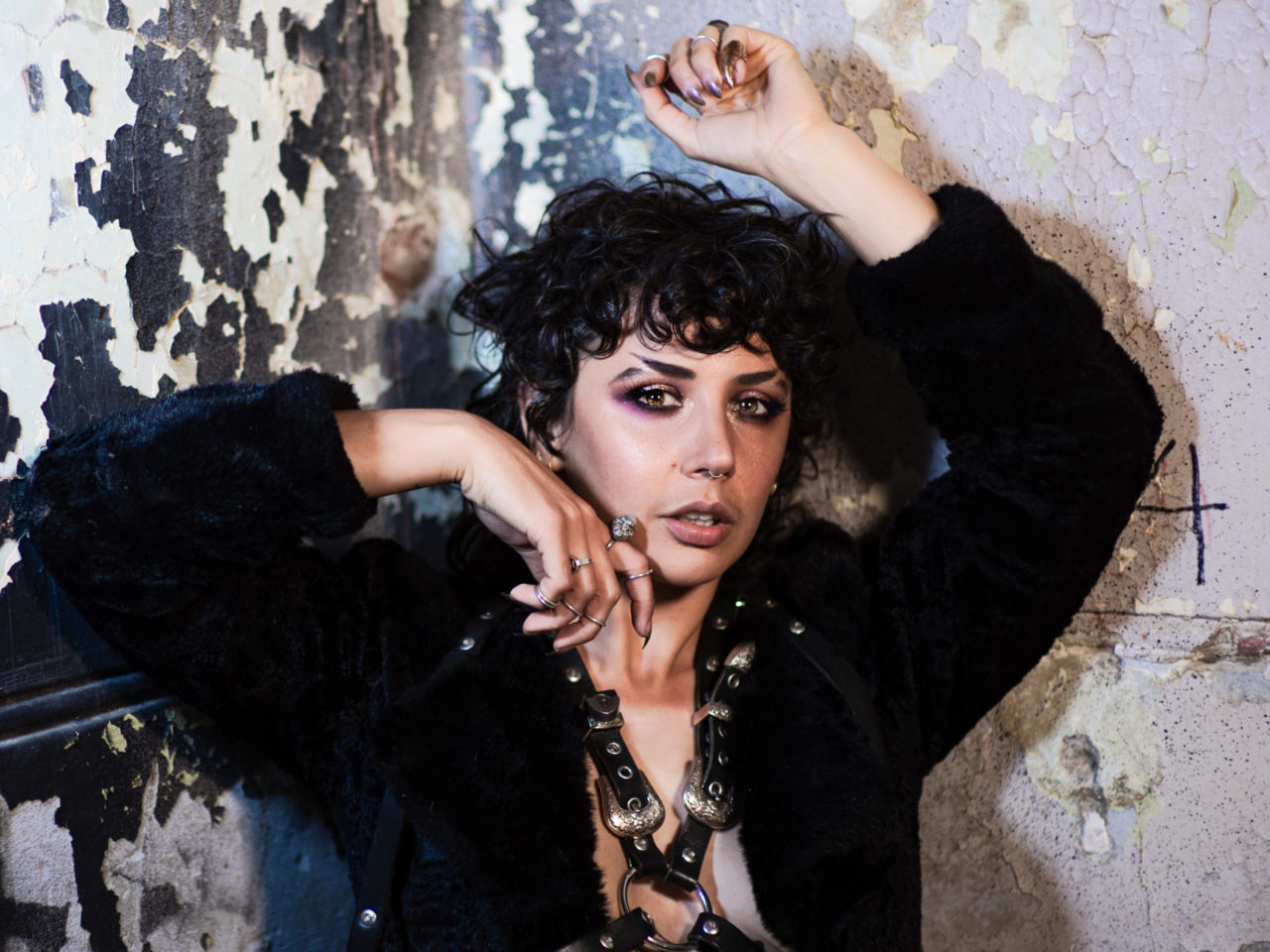 an androgynous, dark featured figure in a black fur coat and leather harness rests against a paint chipped wall with a number 4 scrawled hastily on it.