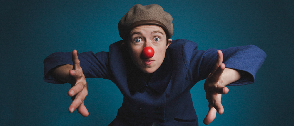 A clown with a red nose holds out hands and looks into the camera
