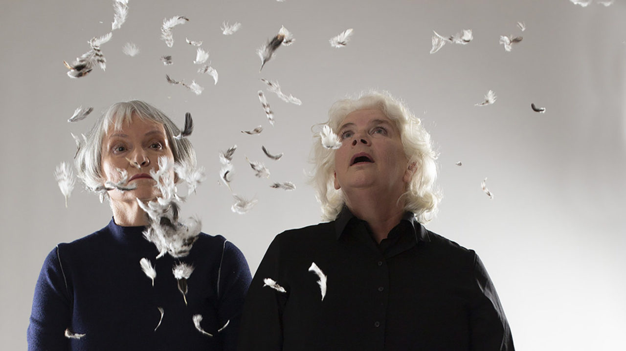 Two women with grey hair look up at feathers floating in the air
