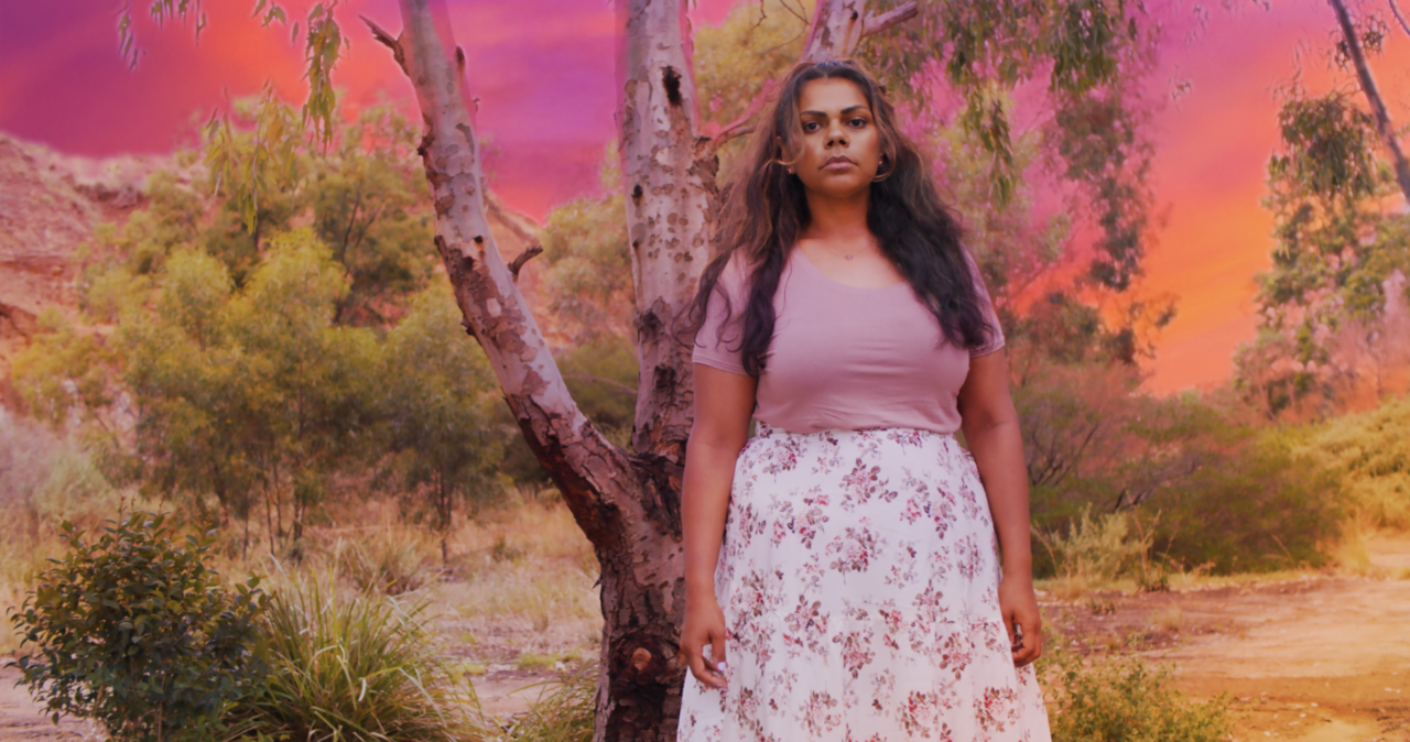 A woman with long dark brown hair and brown skin wears a pastel pink t-shirt and a long white skirt with flowers on it. In the background is a bush landscape with a vibrant pink and orange sunset.
