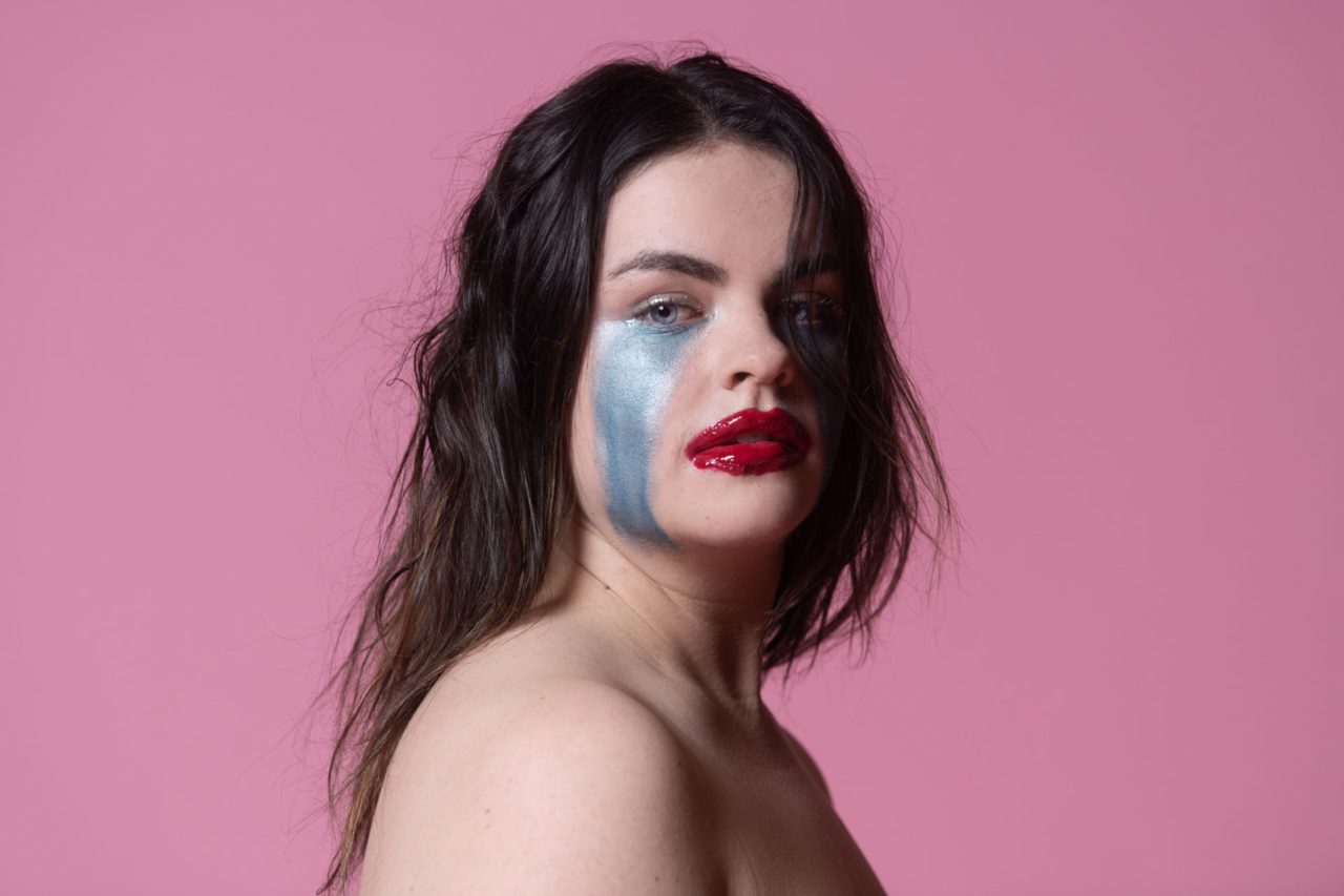 A person with dark hair stares at the camera with a sad expression. She has too much lipstick on, it’s almost clown-like, and blue paint runs down her cheeks like tears.