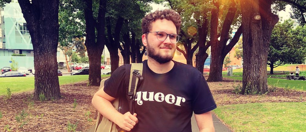 Sam Martin, a young caucasian man wearing black tee shirt with “queer” in white letters and a clear hearing aid, walks through a park