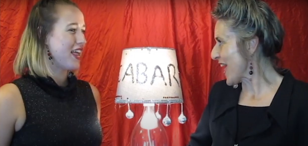 Two woman look at each other with a read curtain behind them and lamp between them