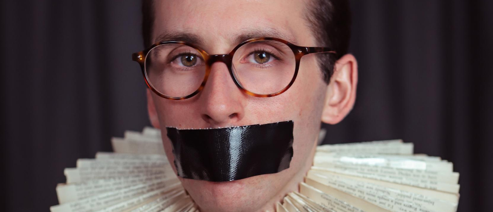 A closeup of a despondent man looking towards the camera, wearing round-framed glasses, a combover, black tape over his mouth, and an Elizabethan-inspired ruff around his neck, made out of recycled book pages.
