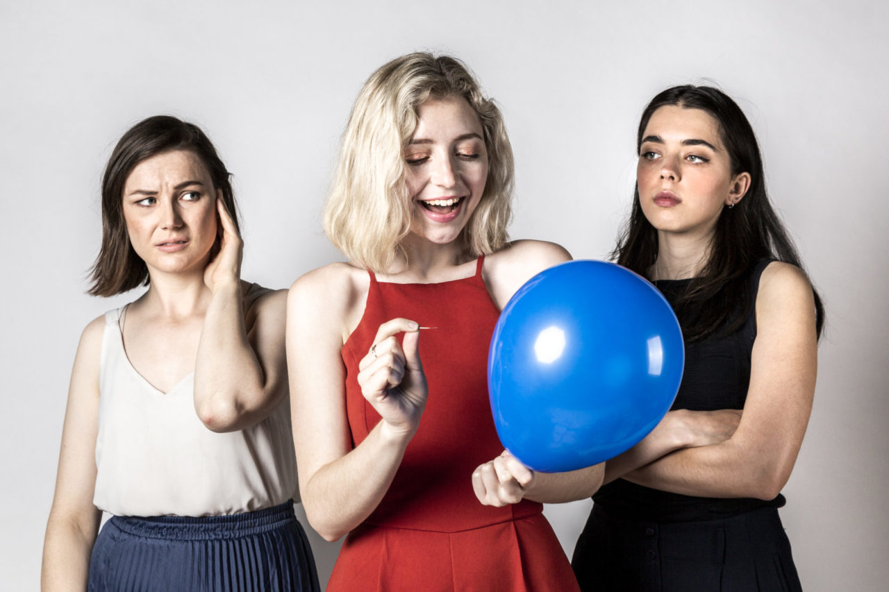 Three women in front of a white photo backdrop. The woman on the left has brown hair and wears a white blouse. She holds her hand up to her ear as she looks with a worried expression at the woman in the middle. The woman in the middle has blonde hair and is dressed in red. She has a big smile on her face, looking at a blue party balloon she is holding which she is about to burst with a pin. The woman on the right has dark hair and wears a black pantsuit with her arms crossed in front of her. She stares challengingly at the woman in the middle.
