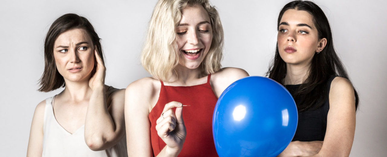 Three women in front of a white photo backdrop. The woman on the left has brown hair and wears a white blouse. She holds her hand up to her ear as she looks with a worried expression at the woman in the middle. The woman in the middle has blonde hair and is dressed in red. She has a big smile on her face, looking at a blue party balloon she is holding which she is about to burst with a pin. The woman on the right has dark hair and wears a black pantsuit with her arms crossed in front of her. She stares challengingly at the woman in the middle.