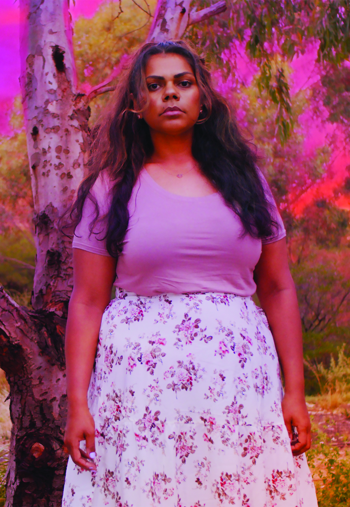 A woman with long dark brown hair and brown skin wears a pastel pink t-shirt and a long white skirt with flowers on it. In the background is a bush landscape with a vibrant pink and orange sunset.