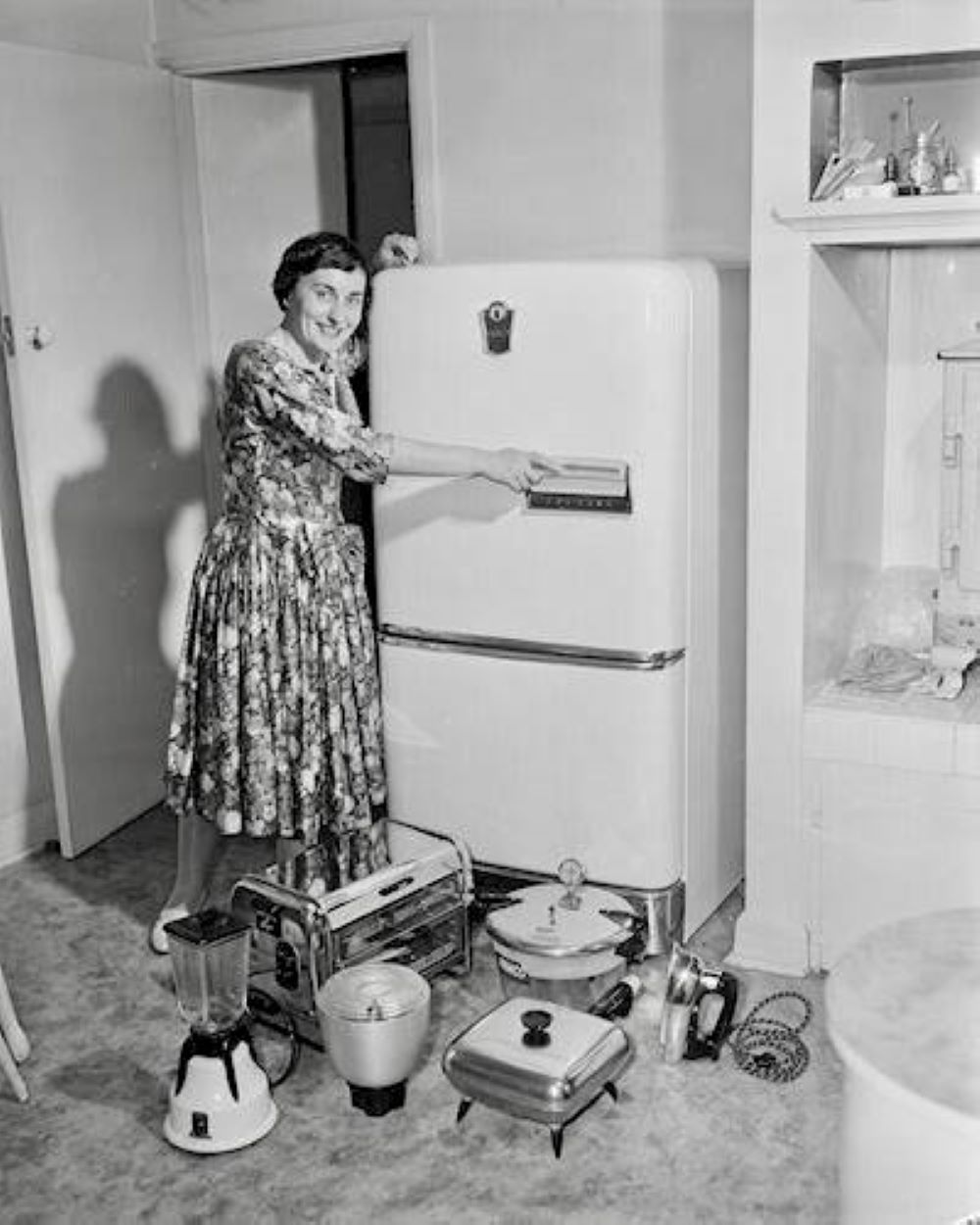 An old photo in black and white of a woman in the kitchen with a long dress pointing at the fridge