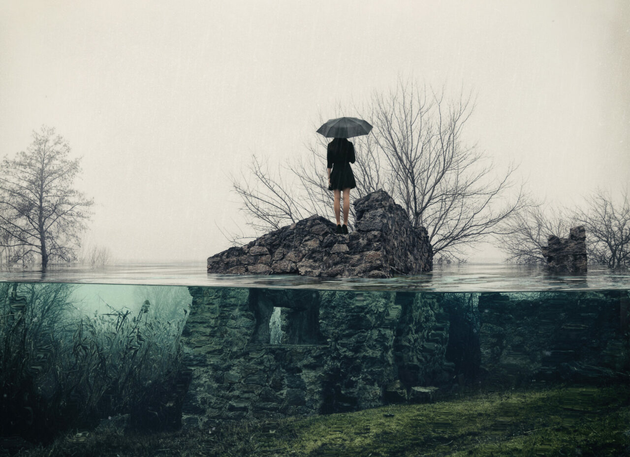 A woman in a black dress stands in the middle of a rocky island with an umbrella.