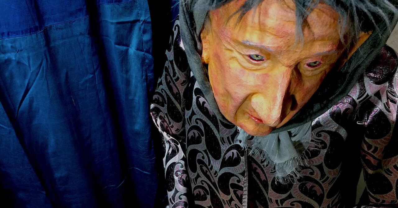 viewed from above, the head and torso of an old lady puppet wearing a patterned jacket, in front of a blue curtain