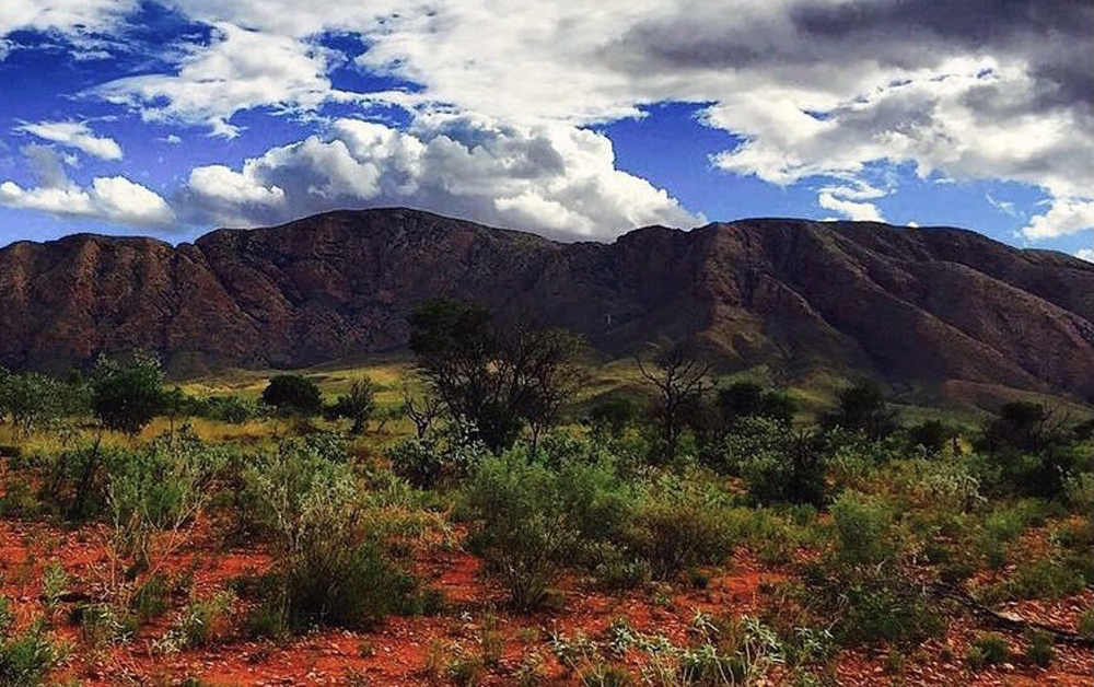 A landscape with red dirt, green shrubs, a mountain and blue sky with clouds.