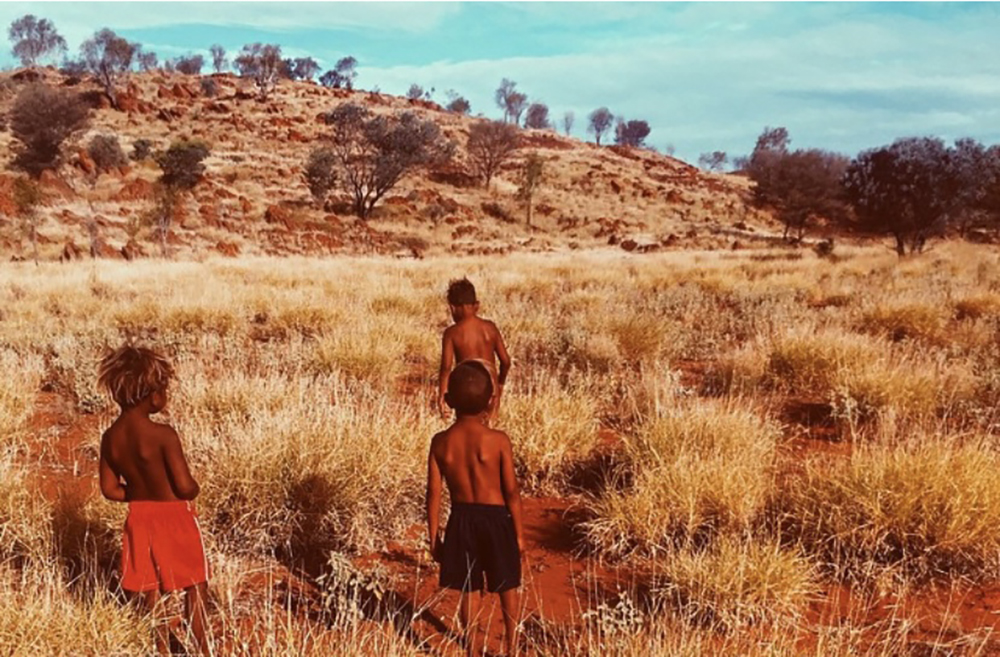 A desert landscape with three young Indigenous children walking towards a mountain through dry grasslands.
