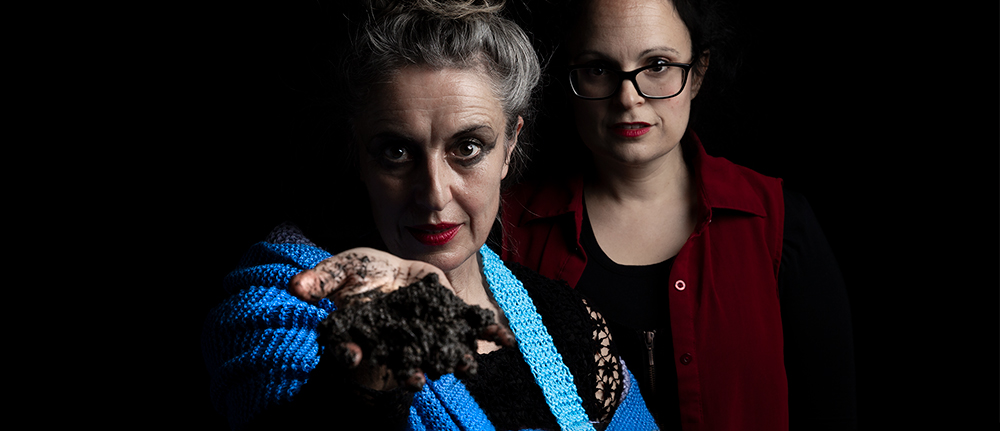 Two women, side by side, looking directly at the viewer. One woman has a coil of knitting in blues, greens, grey curled around her shoulder and wrist, she offers a handful of mud to the viewer.