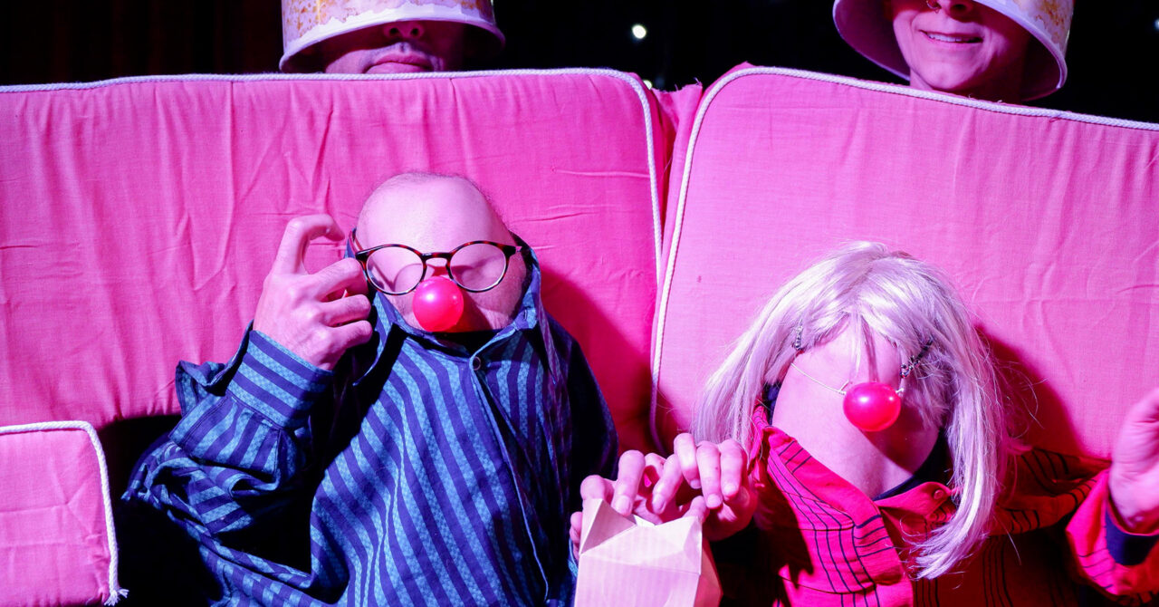 Two puppets, made of people’s knees and dressed in shirts and red noses, sit at the movies eating popcorn.