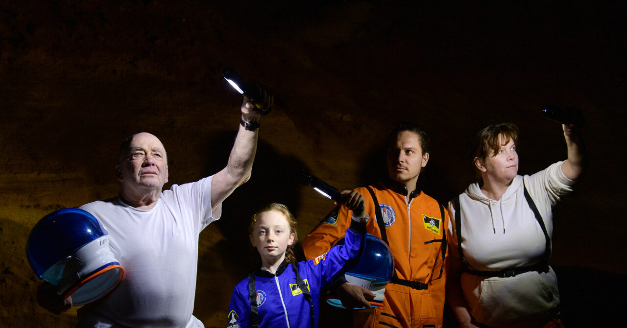 A family — man in his sixties carrying a space helmet, an 11 year old girl wearing a blue space suit, a man in his 30s in an orange space suit carrying a space helmet, and a woman in her 50s stand against an alien looking, rock wall. They hold lights aloft in the darkness.