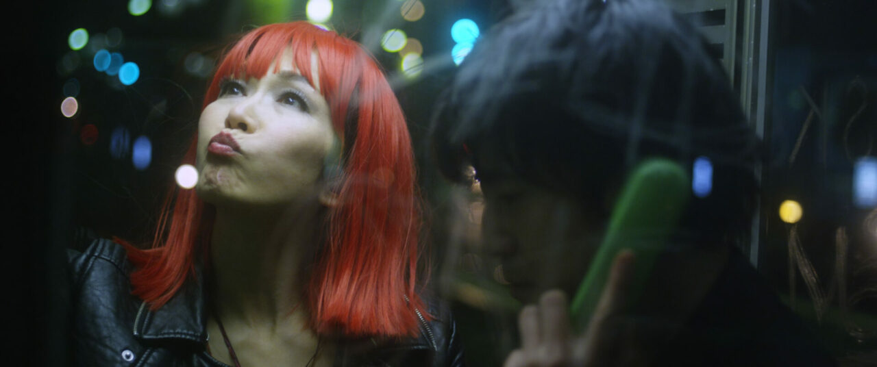 A man and woman in a phone booth at night. The man is on the phone. The woman has red hair and purses her lips.