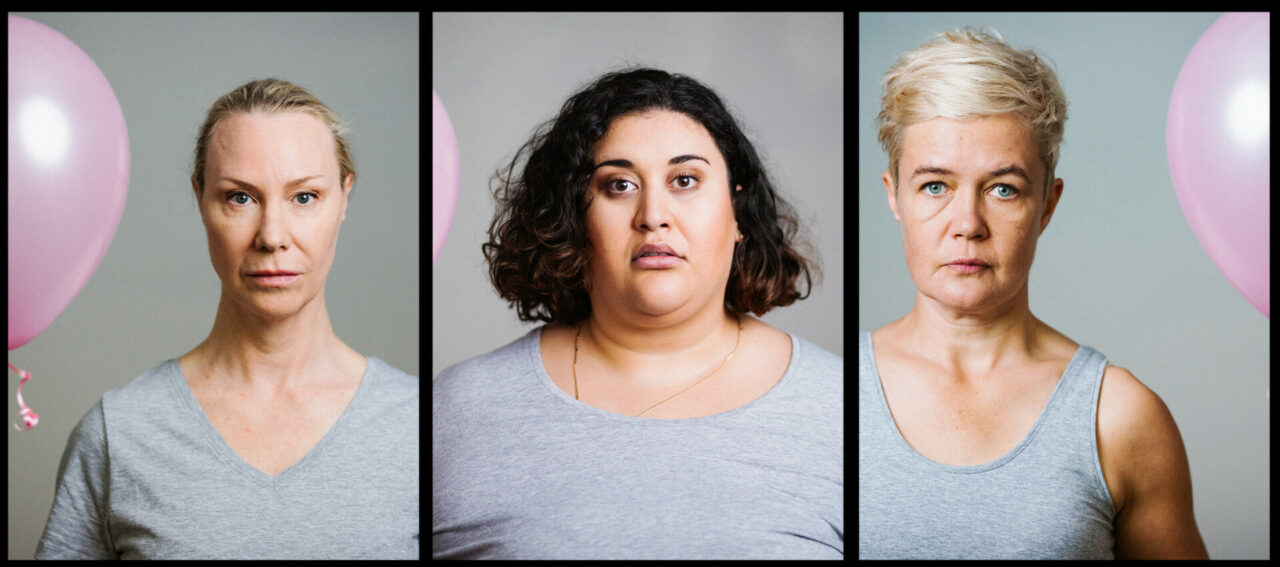Three women in grey t-shirts stand shoulder to shoulder, each holding a pink balloon.