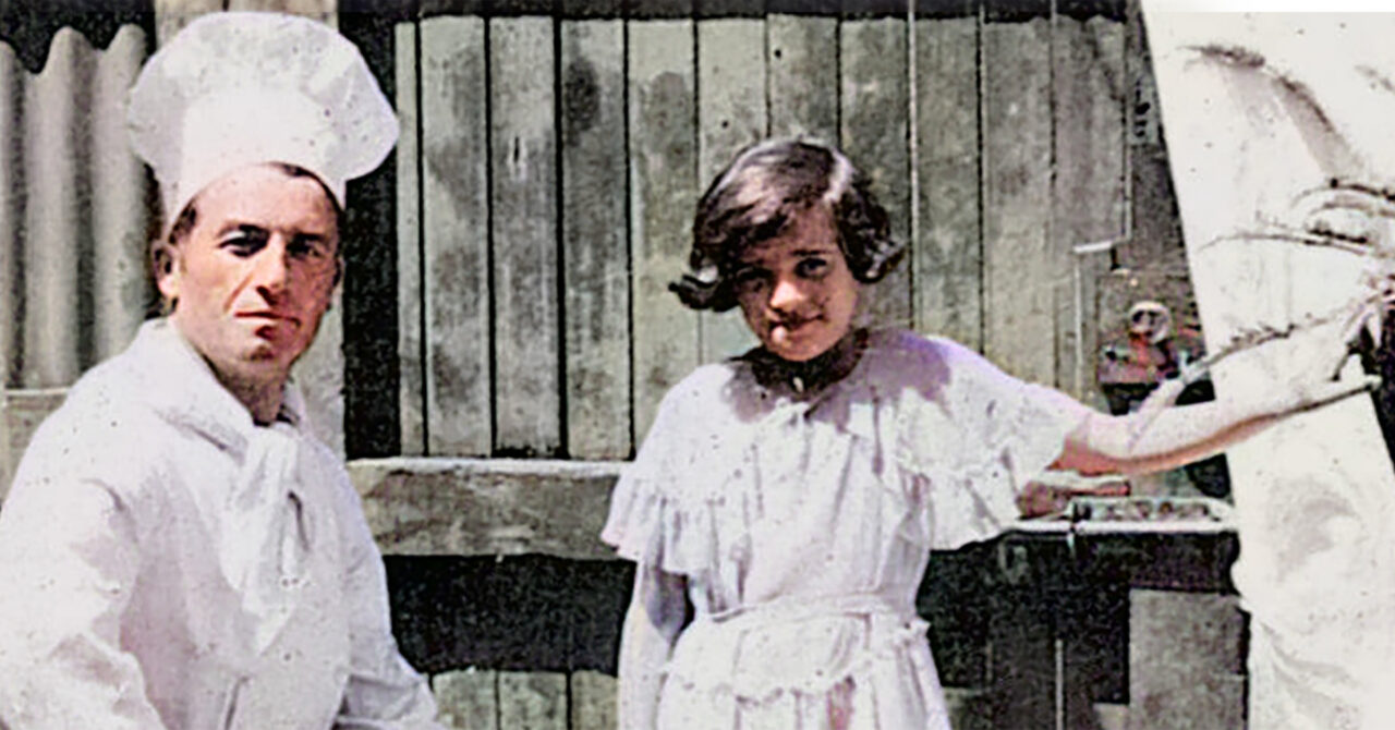 A young girl of 10 years old stands next to a sitting man who is her family’s restaurant’s chef, in front of an old wooden suburban fence with the chimneys of East Melbourne behind her.