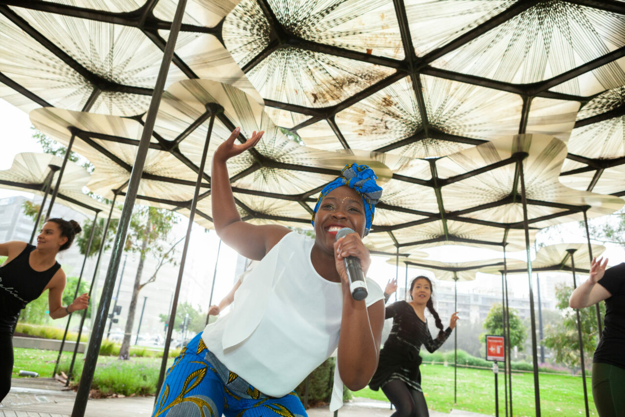 A woman with dark skin smiles and dances with participants behind her as she instructs them.