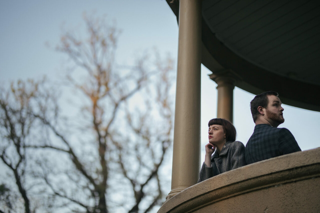 Framed within a large beige gazebo with towering columns stands two people, shoulder to shoulder. The person closest stands with his back facing out and he is a white man wearing a checked grey suit jacket, his gaze is up and out into the distance. Next to him is a white woman facing outwards and staring up at the horizon line. She is wearing a fur-like coft and her hand is curled underneath her chin. To the lest of the gazebo is a large tree with just the upper branches catching the warm golden sunrise on its limbs.
