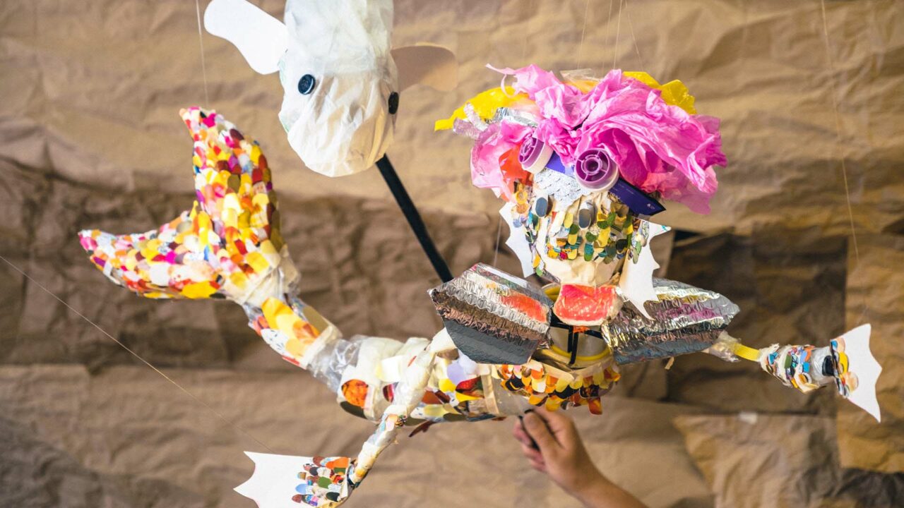 A brightly coloured Mermaid puppet is swimming below a white Porpoise puppet. Both puppets are made from recycled rubbish.