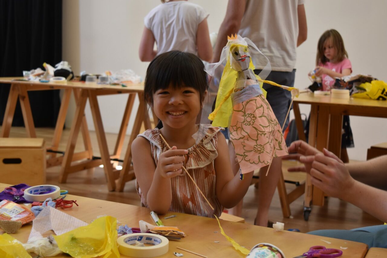 A young girl holding up a puppet she made herself. She has dark hair with a straight fringe. She smiles widely.
