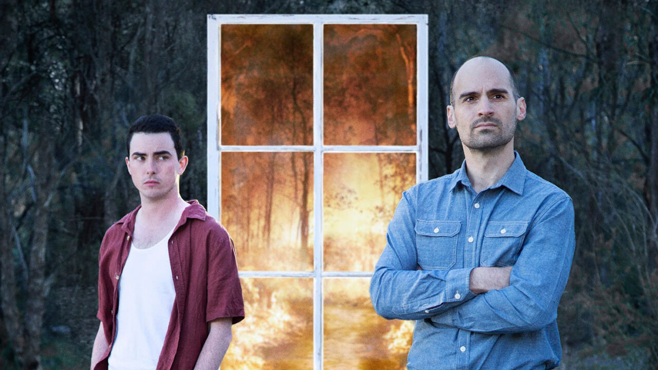 Two brothers standing in the bush, one with hands in his pockets, the other with his arms crossed. In the background is a view of a bushfire through a window.