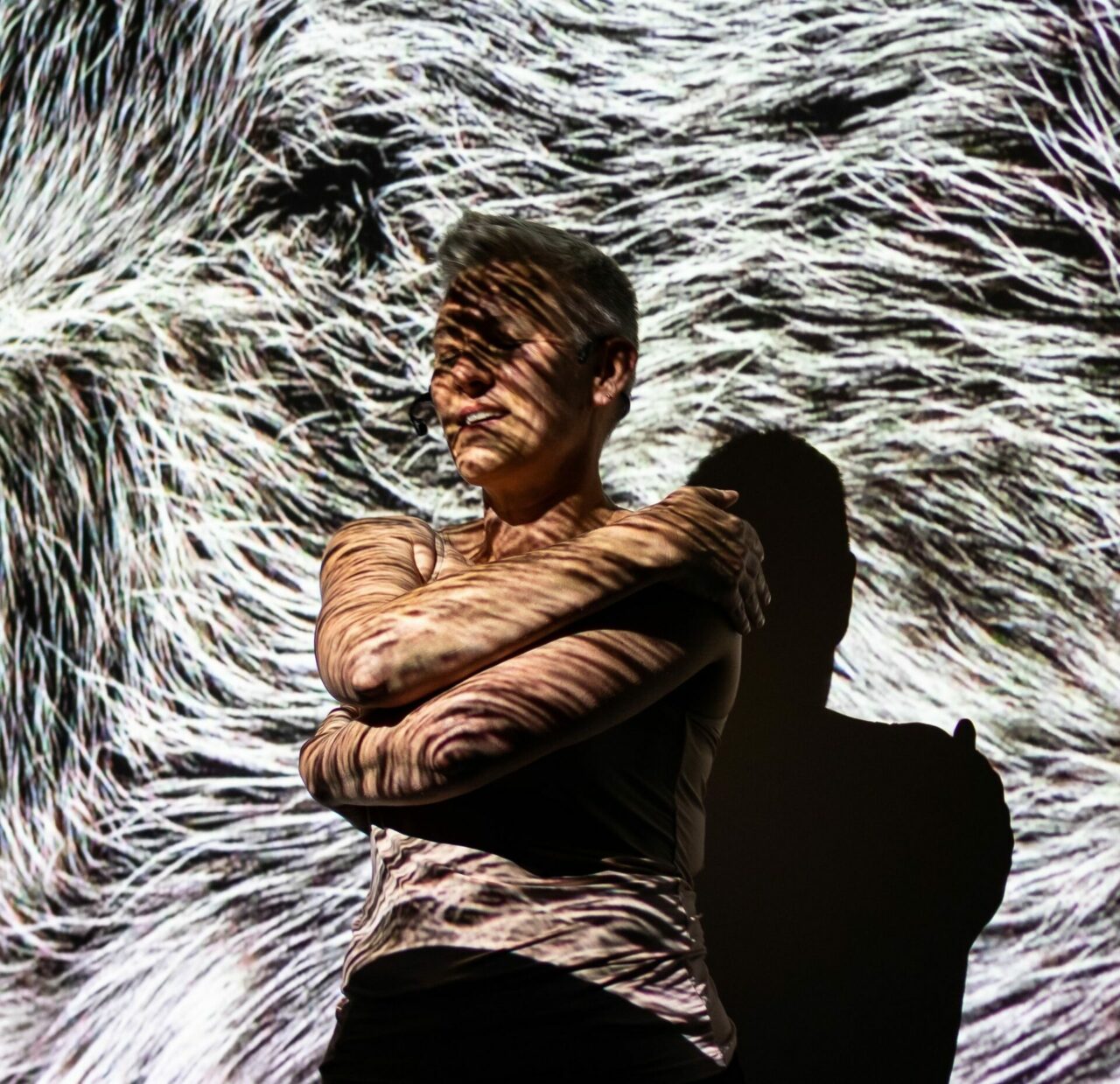 Woman with short blonde hair and eyes closed, hugging herself in front of an animal fur projection.