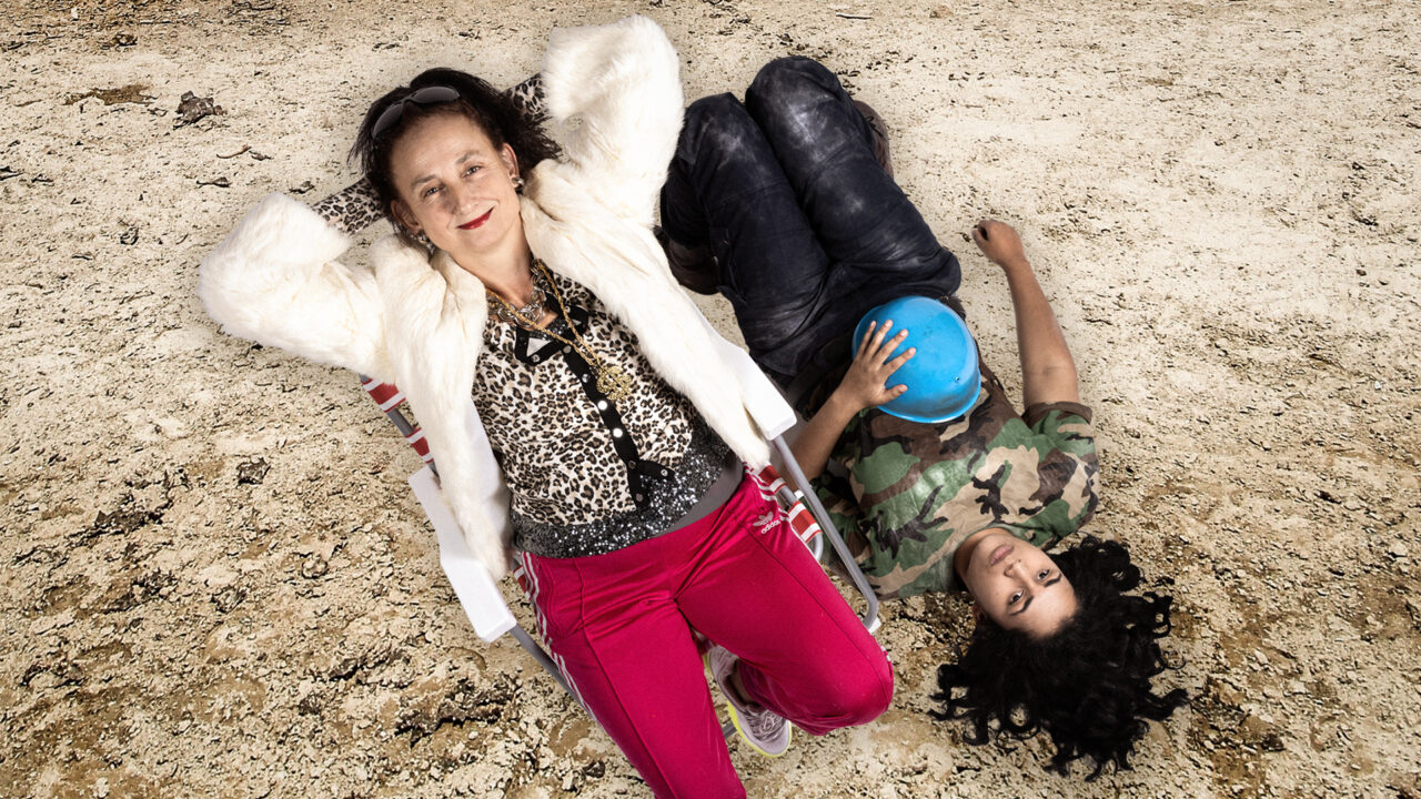 A smiling woman in gold jewellery reclines on a chair, beside a younger woman in camouflage lying on the dusty ground.