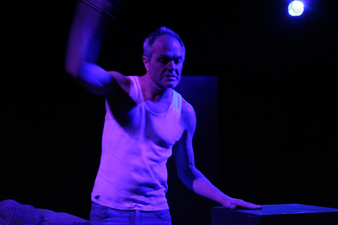 An older man in shabby clothes, standing in dark purple light, stretches one arm up above his head.