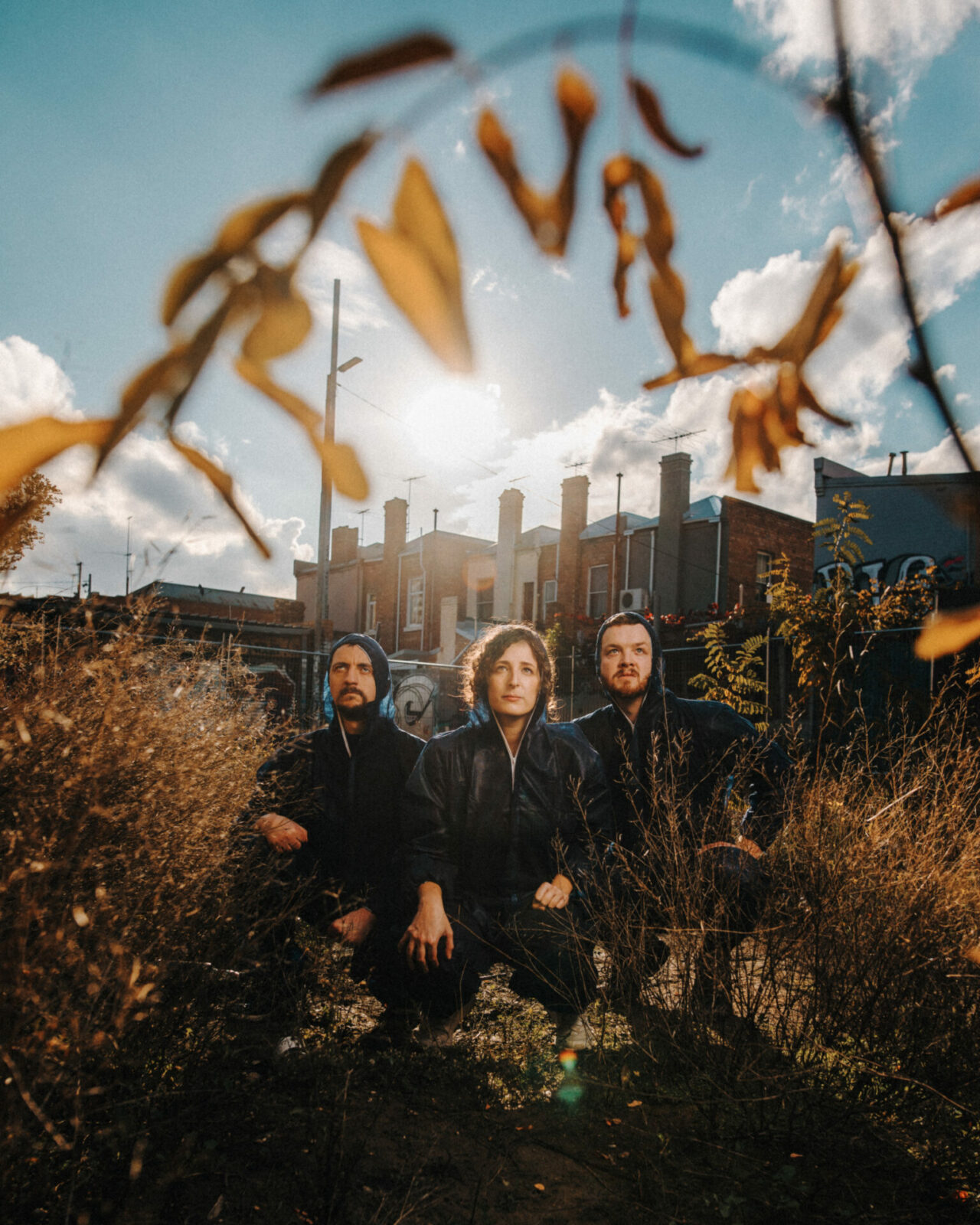 three people are squatting in a decrepit landscape, dying trees in the foreground and old buildings behind them. Golden light shines from behind as they stare up past camera, determination on their faces.