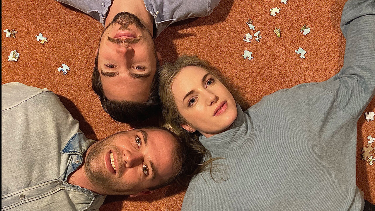 A woman and two men lie with their heads together looking up, there are puzzle pieces scattered around them.