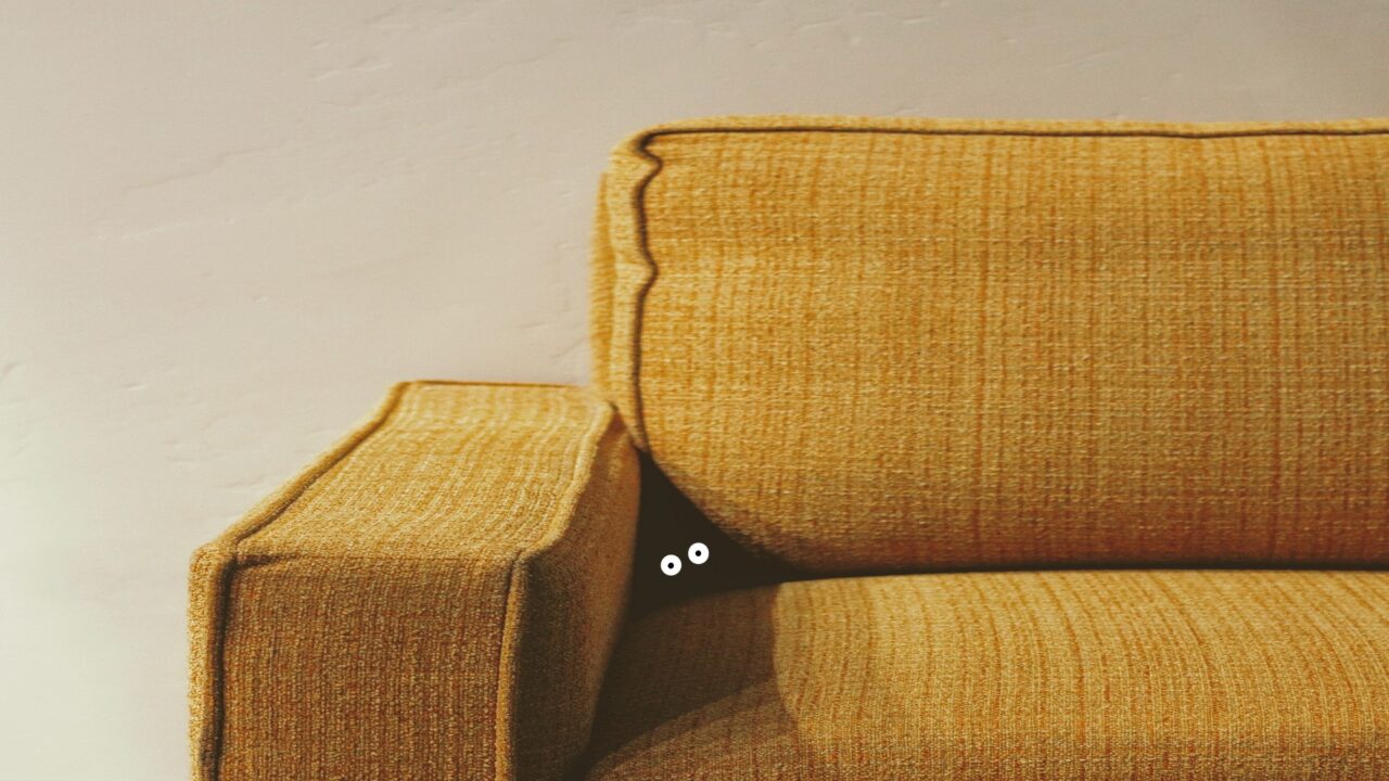 A pair of white cartoon-like eyes stare out from the shadowy crevice of a yellow couch. The couch sits against a plain cream-coloured wall.