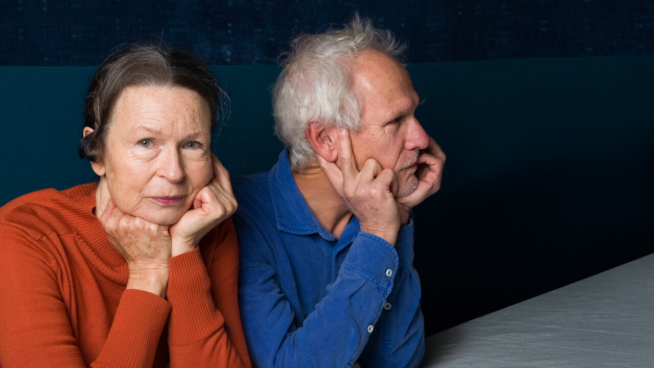 A woman in an orange shirt sits at a table beside a man in a blue shirt. She looks at the camera, he is in profile.