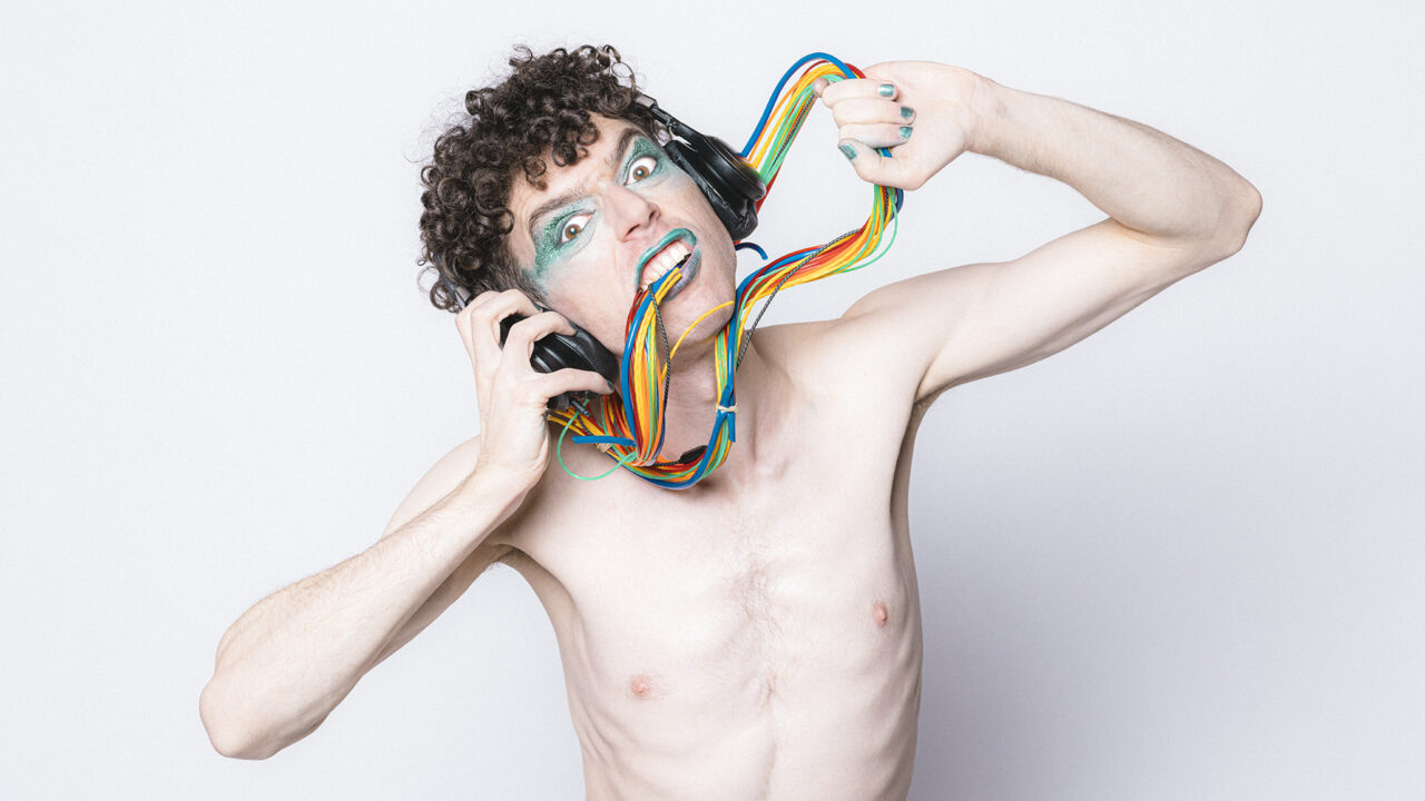 A person with curly hair and glittery makeup wears headphones with computer wires tangled around their neck and in their mouth.