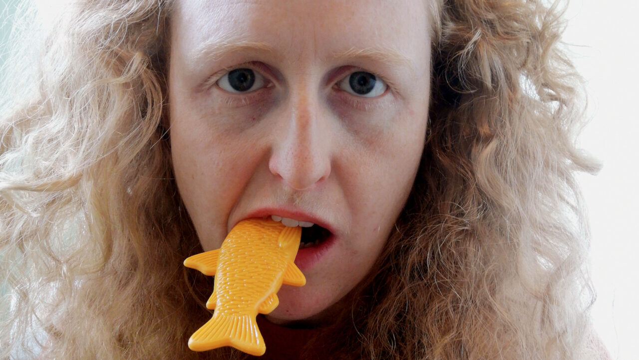 A woman with white skin and blonde curly hair stares into the camera with the tail of an orange plastic toy fish sticking out of her mouth.