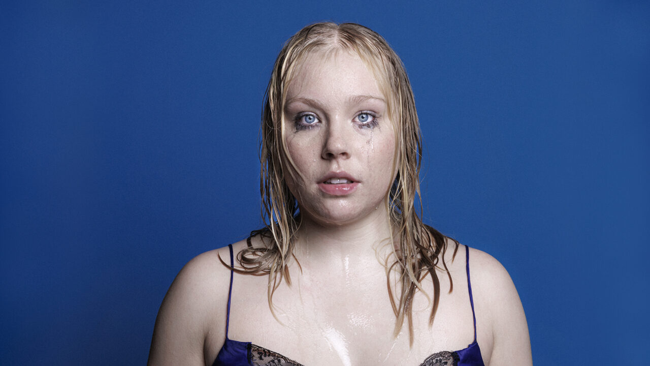 A blonde woman in a blue dress stands dripping wet on a blue background, looking directly at the camera.