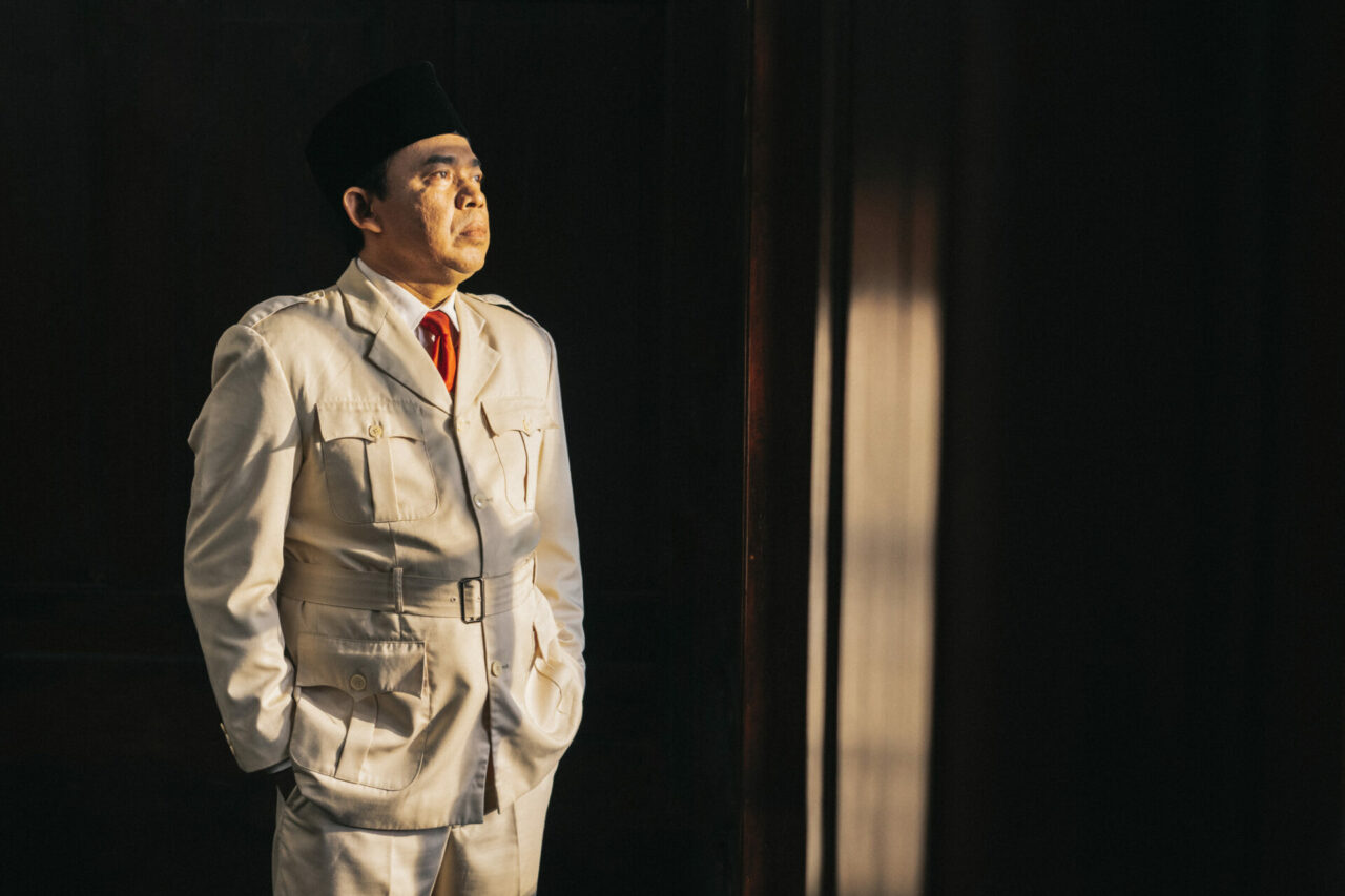 A man in a broken-white suit stands in the front of a chair, he’s wearing a black hat.
