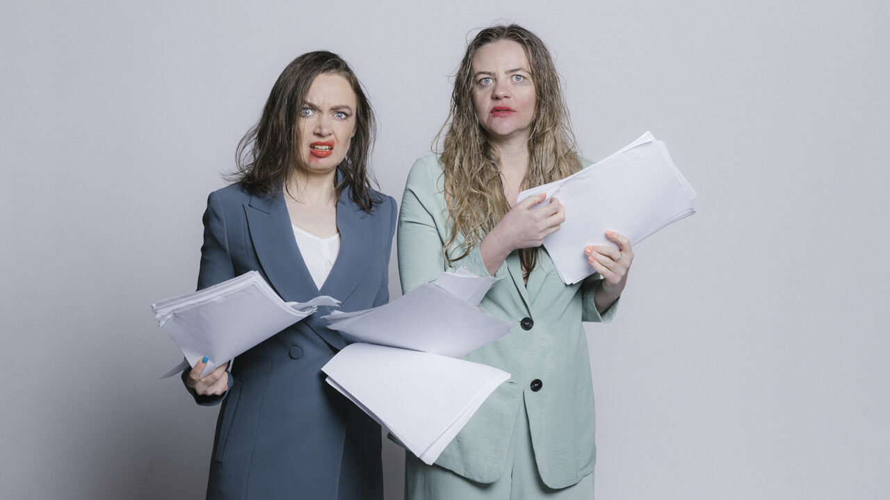 Two women in business suits stare into the camera with horrified looks on their faces. They clutch reams of paper, their hair is wet and their make-up smudged.