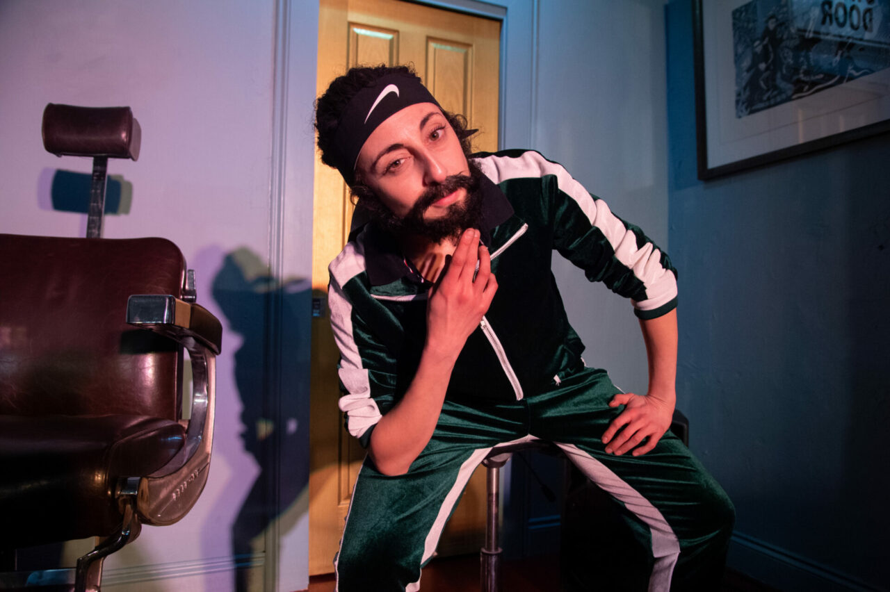 A woman of Arabic descent, wearing a beard and a dark green matching tracksuit smiling cheekily at the viewer.